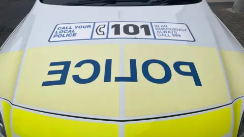 The bonnet of a yellow and white police car with the word 'police' written in large blue block capitals backwards. The bonnet has a sticker on it telling people to call 101 or 999.