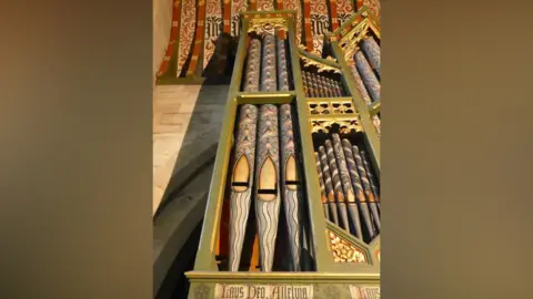 Bridget Weaving Green-painted casing around black and gold pipework on a 19th Century organ in a church. Just the top of the pipes can be seen, stretching up to a red and black painted ceiling.