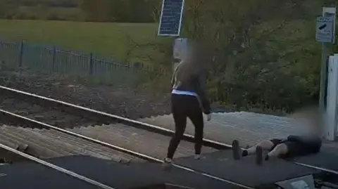 A person in shorts with their face blurred is seen lying on a railway track. A person in black trousers and a jacket with their face blurred is walking over the track towards them.