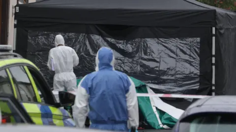 Pacemaker Two forensic investigators in hazmat suits walk towards a temporary black tent which has been erected at the scene where a man's body was found in a burning car in Portadown. A yellow and blue police car is parked close to the tent and a red and white police cordon is stretched across the tent.