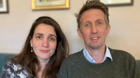 BBC Andrew Davies (right) and his wife Milica. They are sitting on a green sofa and pictured from the chest up. She has long dark hair, is wearing a black and white marble-effect dress and drop pearl earrings. He has on a blue shirt with the collar unbuttoned and a dark green jumper over the top.