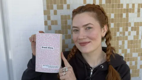 Writer and journalist Kate Pasola holding her book Bread Alone. She has red hair and is wearing a black hoodie. The book is white with red writing on the cover.