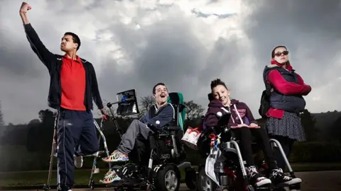 Four people outside, with one person on the left standing using sticks and raising a fist. Two others are seated in wheelchairs, one of which has a communication device attached, and another is stood with their arms crossed and wearing sunglasses. A cloudy sky and trees are visible in the background.