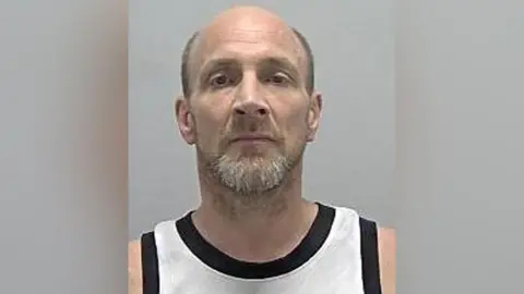 A custody image of Matthew Haydon, he is looking straight at the camera, wearing and black and white to top. He has a beard and short hair. 