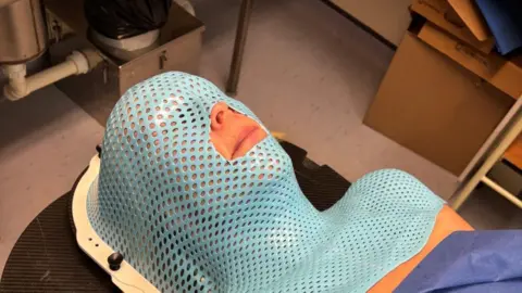 Margot lying on a medical bed wearing a blue mask perforated with hundreds of holes covering her face and upper chest