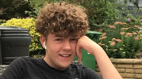 Thames Valley Police A close-up of a teenage boy leaning on his hand and smiling, with a headphone in his ear, and bushes in the background 