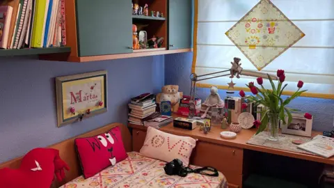 A small bedroom contains a picture hanging on a wall with the word "Marta" and a pillow on a bed with the word "love". A vase of flowers stands on a desk alongside books, photographs and ornaments.