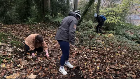 People earthworm sampling in Colin Glen. There is someone on the left in a brown top with brown hair, a person in a grey jacket with blue jeans and black and white striped hat in the centre and someone in black clothing in the distance, far right, with blue bag on their back. They are hunting among leaves.