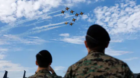 SeongJoon Cho/Bloomberg via Getty Images Eight black and orange military jets swoop over the heads of two soldiers wearing camouflage fatigues and berets. The sky is blue with a few clouds.