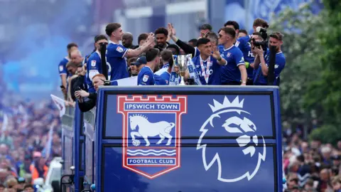 Action Images/Reuters Ipswich players all wearing blue and standing on the top deck of an open top bus. There is a trophy at the front. The bus is blue with the Ipswich Town and Premier League crests on it, and is surrounded by a large crowd on the floor.