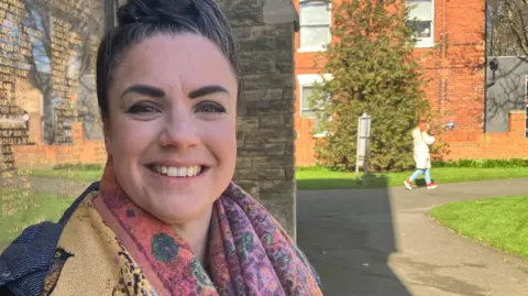 A head and shoulders photo of Katie-Lou Green smiling at the camera. She is wearing a black coat and a multi-coloured scarf. She has black hair which is tied up. She is stood outside in front of a stone building and memorial plaque. 