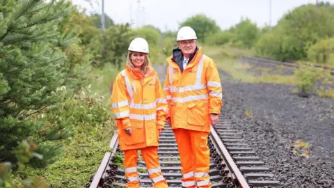 BBC A blonde woman standing next to an older man. Both are wearing orange hi-vis jackets and trousers and hard hats. They're standing on a railway line surrounded by green bushes.