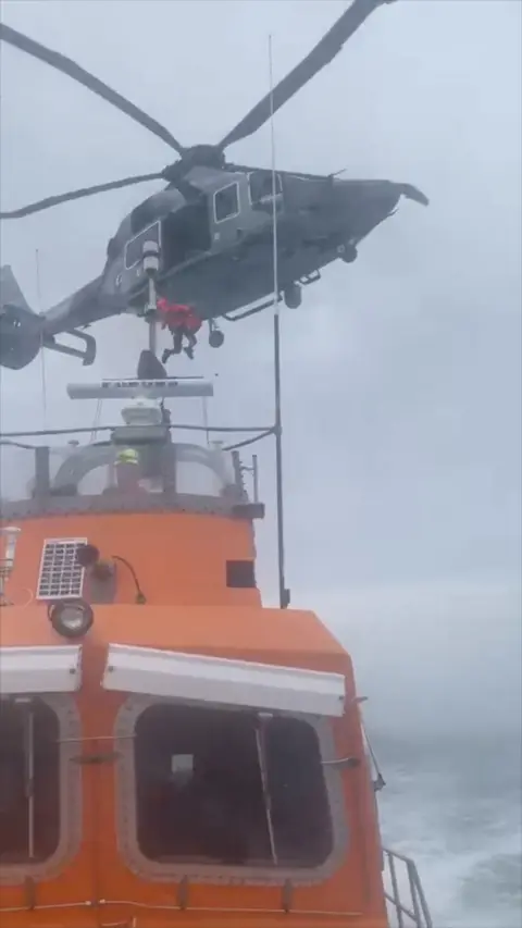 An orange lifeboat sailing with a grey helicopter flying above 