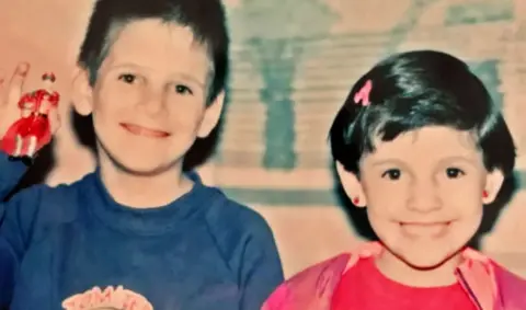 A boy in a blue top and a girl in a pink top, both with dark hair, smile at the camera - the boy is holding up a toy figurine 