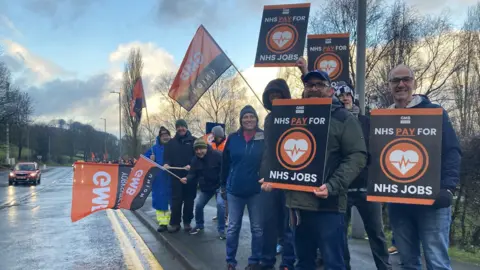 CHARLES HESLETT/BBC Workers on a picket line at the side of a road in Bradford. They are holding black and orange placards which say, "NHS pay for NHS jobs". Some people in the background are holding GMB flags. There is a red car on the other side of the road.