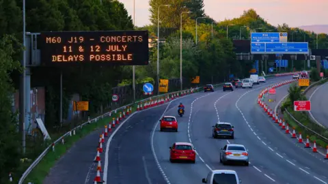 Emma Gibbs A motorway sign above a carriageway, which has a few cars driving past into the distance, reads: M60 J19 - Concerts 11 & 12 July Delays Possible