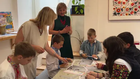 Teachers and pupils at a Ukrainian Saturday school look at work they are doing on a table. There are two female adults, four boys and a girl.