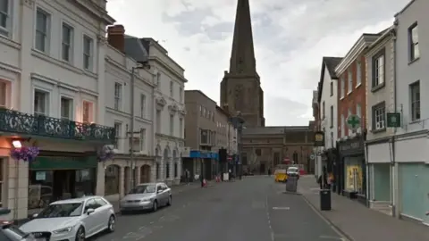 The image shows Broad Street in Hereford. Some cars are parked on the left outsides shops/businesses and there are some empty shops on the right hand side. The street is wide and most building are white/cream in colour. A church tower/spire can be seen at the end of the street. 