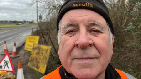 John Devine/BBC Mark Fishpool, 68, has grey hair hair around his ears, he is wearing a black beany hat with a high-viz orange jacket. Behind him are a selection of old road signs, stands for them, bollards, sandbags and safety lamps.