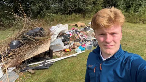 A man with short strawberry-blonde hair wearing a blue rain jackets stands beside a large pile of rubbish