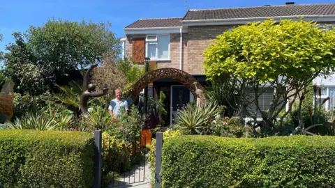 Katie Jaffar Steve Damon is pictured standing in his front garden. He is surrounded by tall trees and shrubbery and there is a bronze statue of a figure with arms outstretched beside him. There is a rusted bronze arch on an angle on the other side of him. The arch is over a pathway which leads to his front door. A neat front hedge and a black metal gate can be seen in front of the garden.