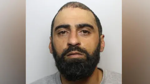 A police mugshot of Sarmad Khawaja, who has a black beard, dark eyes and short, shaved black hair.