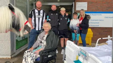 Allison Charlton, an elderly woman, sits in a wheelchair and looks on as a horse called Charlie stands to her side. A hospital bed is to her right and it is outside of a building entrance. Two men and two boys and one girl stand behind the woman.