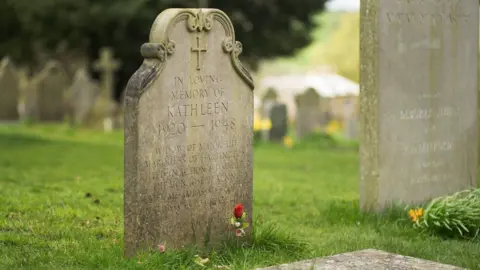 Getty Images A weathered gravestone stands on a neat grass lawn in a cemetery, with other headstones in the background. Inscribed on the stone are the words: "In Loving Memory of Kathleen 1920 to 1948". A red flower grows at its base.