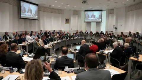 John Fairhall/BBC A large council room filled with men and women all sitting on chairs and in front of tables in the round.