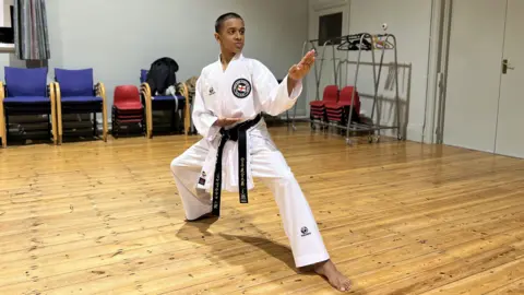 Mayon stands in a karate stance with his hands outstretched, wearing a traditional gi with black belt and club badge. In his standing on a wooden floor in a dojo.