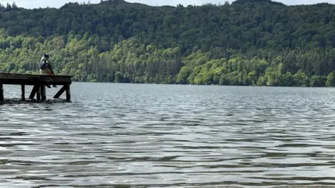A general view of Windermere. A person is sat on the edge of a small pier looking out to the water. On the other side of the lake, the shore is green and covered in trees.
