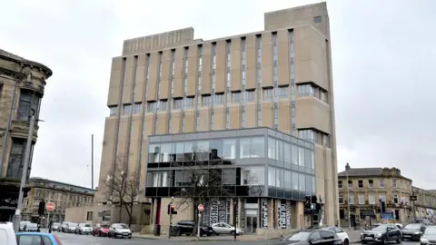 A large, multi‑storey concrete building on a street corner in an urban area. The building has a distinctive, Brutalist-style design, with tall vertical concrete panels running down the front. 