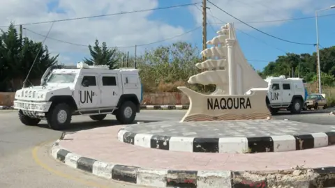 Getty Images Two UNIFIL armoured vehicles outside the entrance of the southern Lebanese town of Naqoura. They are parked beside a roundabout with a statue with the word Naqoura on it. There are blue skies in the background. 