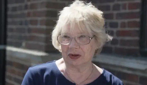 A lady with short fair hair and glasses. She is standing in the street in front of a building.