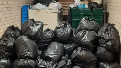 Kay Friday Piles of black bin bags next to two large bins which are overflowing with rubbish.