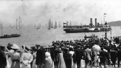 Getty Images Black and white archive image of crowds in Edwardian-era clothing looking out to sea where there are yachts in the distance with sails and flags, and a steam ship