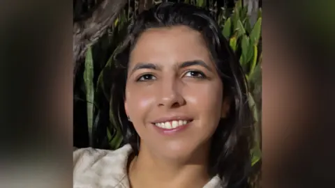 Essex Police Vitoria Figueiredo Barreto is smiling at the camera. She is wearing a light coloured shirt and there is greenery behind her. She has dark coloured eyes and hair. 