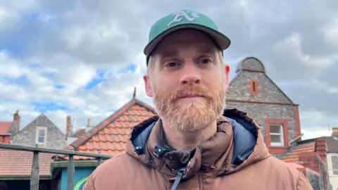 Claire Cavanagh A picture of parent Huw Illingworth standing outside Fairlawn School in Bristol wearing a brown anorak. He has a beard and is wearing a cap and is in front of school buildings and an iron fence. 