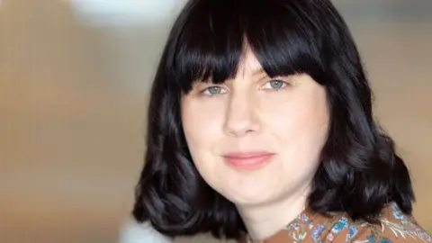 ACE Bonnier Dr Ceri Houlbrook, who has shoulder-length black hair with a long fringe. She is wearing a just-glimpsed brown top with blue and white flowers and leaves and is looking over her shoulder with the beginnings of a smile 