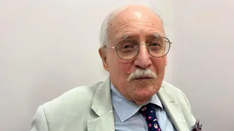 An elderly man is smiling at the camera. He has a white moustache and rounded framed glasses. He is wearing a cream blazer, pale blue shirt and navy blue and pink polka dot tie. 