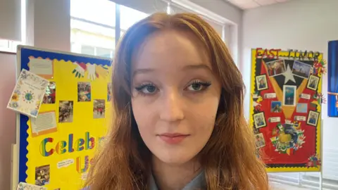 A teenage girl is standing in school. She has long auburn hair. There our board with art on them behind her. She's wearing her school uniform.