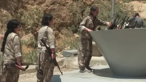 Reuters Three women in light green uniforms and dark hair line up carrying their guns beside a large cauldron