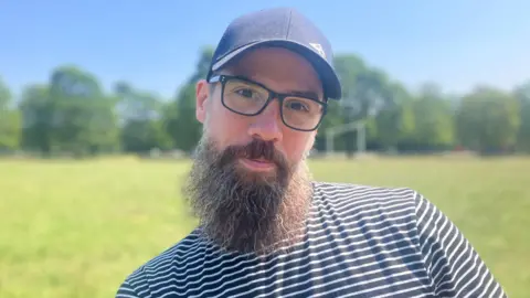 A man with a beard wears glasses and a cap. He has a striped blue and white shirt on. Behind him green space is blurred