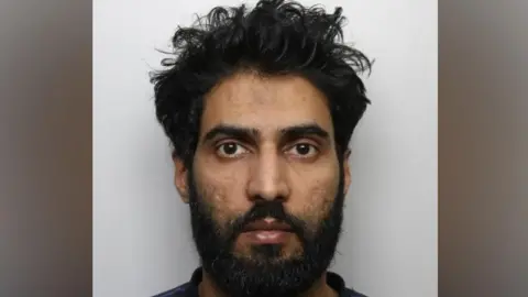 The image is police-style custody or mugshot photograph taken against a plain light-grey background. The picture shows a person facing directly towards the camera, with dark, curly hair and a full beard and moustache. 