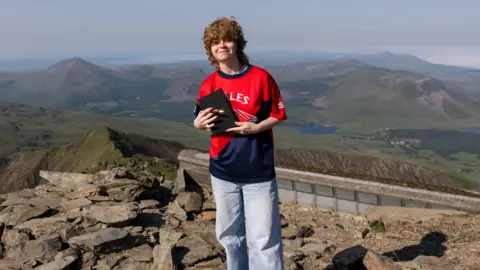 FAW A woman with curly red hair, wearing a red Wales top and blue baggy jeans and holding a black notebook. She is stood on rocky terrain with a mountain landscape behind her.