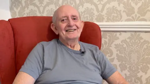 David Rigg is sitting in a red armchair and smiling brightly at the camera. He is bald on top with grey hair at the sides and is wearing a grey t-shirt.