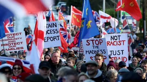Reuters A large crowd gathers at a protest in Crowborough carrying Union flags and banners. Visible signs include messages reading "Stop the Madness" and "Not Racist". People fill the street in the foreground and background.