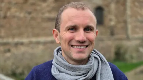 Stuart Woodward/BBC Councillor Lee Scordis stands in front of Colchester Castle. He wears a blue jumper with a grey scarf around his neck. He has close-cropped brown hair and stubble, and is smiling at the camera