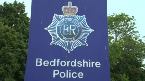 The blue sign outside Bedfordshire Police Headquarters. It bears the troop's coat of arms and the lettering "Bedfordshire Police". There are trees behind the sign.