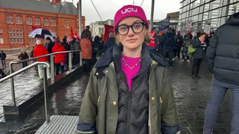 Jo Grady outside the Senedd in pink UCU hat, matching pink jumper over which she is wearing a black puffer jacket and a green wax jacket. She wears glasses.